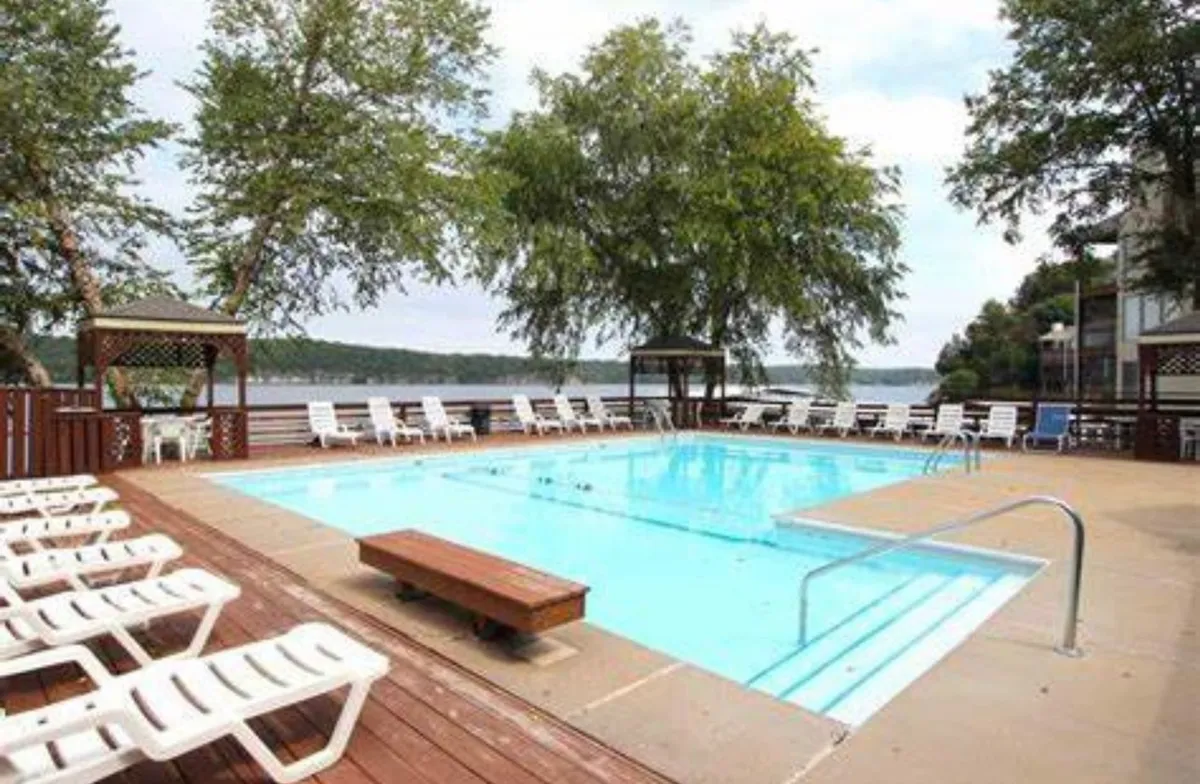 The scene features a serene swimming pool surrounded by lounge chairs, a gazebo, mature trees, and a glistening lake in the distance. The environment evokes feelings of privacy, relaxation, and a connection to the natural surroundings.