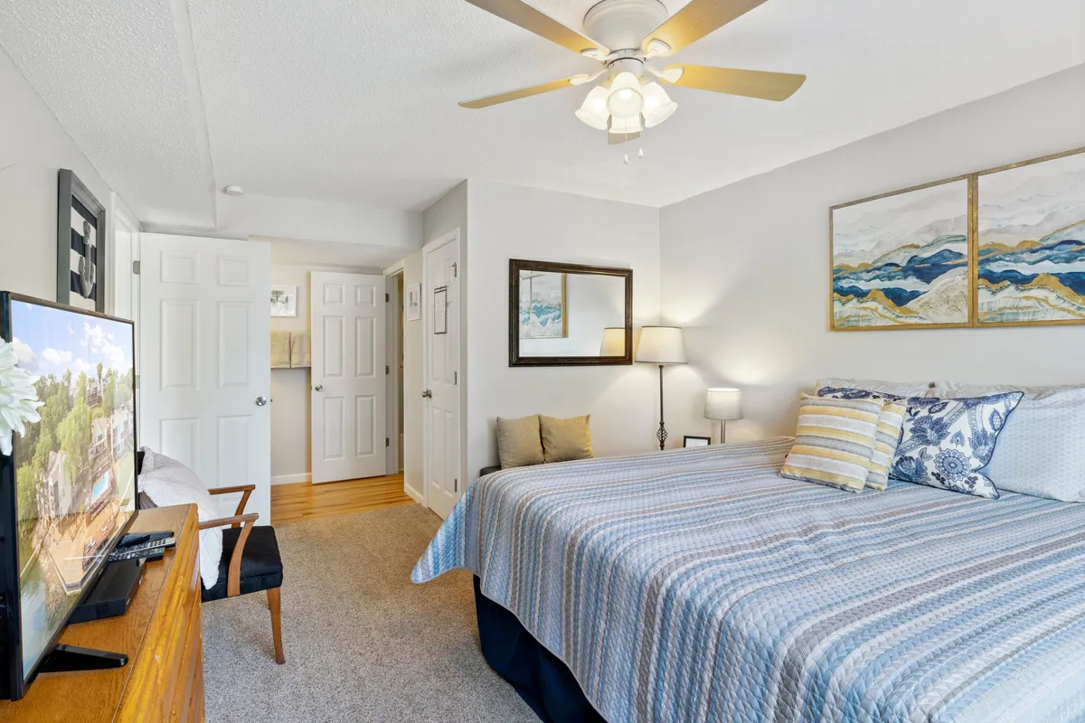 A serene bedroom with a blue bedspread, abstract wall art, a television atop a dresser, and a bedside lamp. Natural sunlight streams in, enhancing the room’s tranquil and restful vibe.