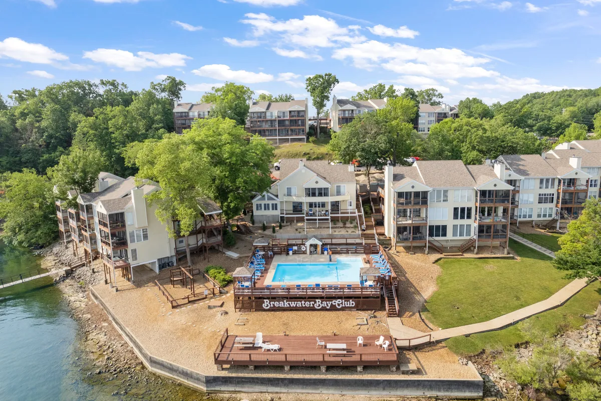 This exterior image showcases a resort-style swimming pool surrounded by lush trees and set against the backdrop of the waterfront and clubhouse. The atmosphere is relaxed and inviting, with plenty of space for swimming, sunbathing, and gathering for social activities.