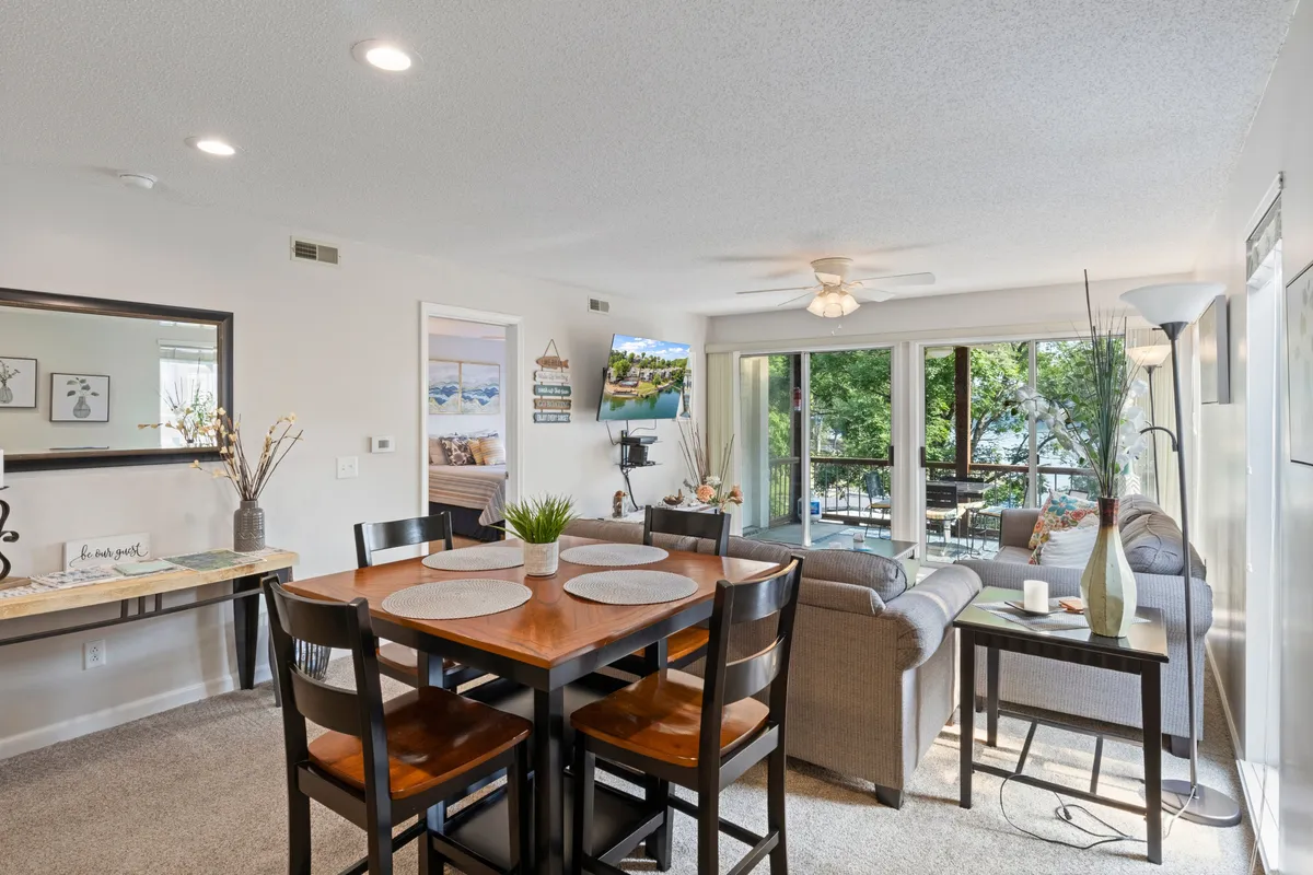 A cozy dining and living area featuring a dining table set near large windows, a comfortable sofa, decorative plants, and colorful wall art. The space is filled with natural light, making it ideal for social gatherings, family meals, or quiet breakfasts.