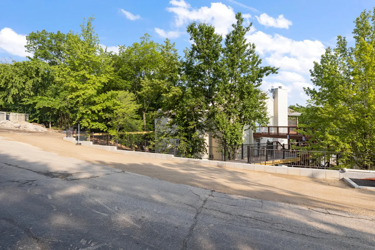This exterior image highlights the lush greenery surrounding the property, with a driveway leading up to a modern building. Trees frame the setting, offering a tranquil and secure atmosphere ideal for peaceful mornings or unwinding after work. The contemporary architecture and foliage suggest a welcoming environment well connected to nature.