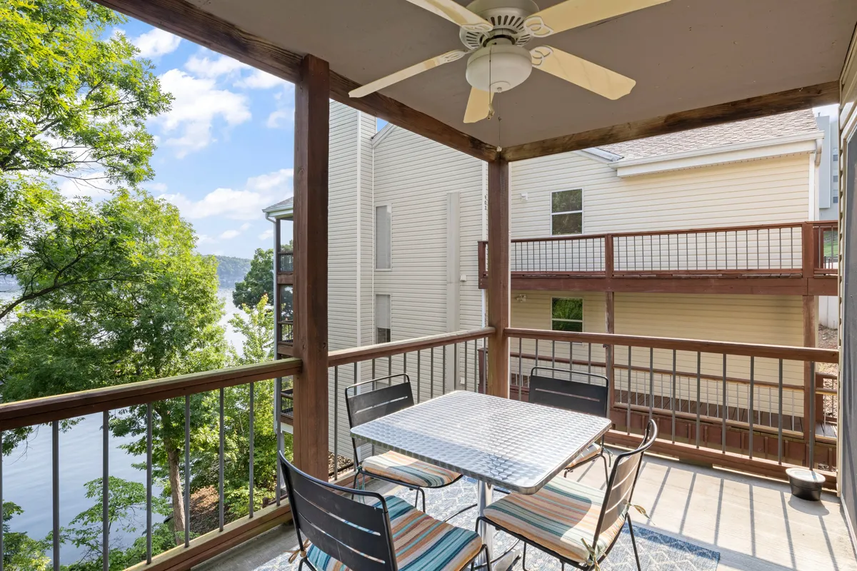 An outdoor dining table sits beneath a ceiling fan on a covered balcony or patio, offering panoramic views of the lake. The relaxed setup is perfect for enjoying morning coffee, sunset dinners, or evening chats with friends and family.