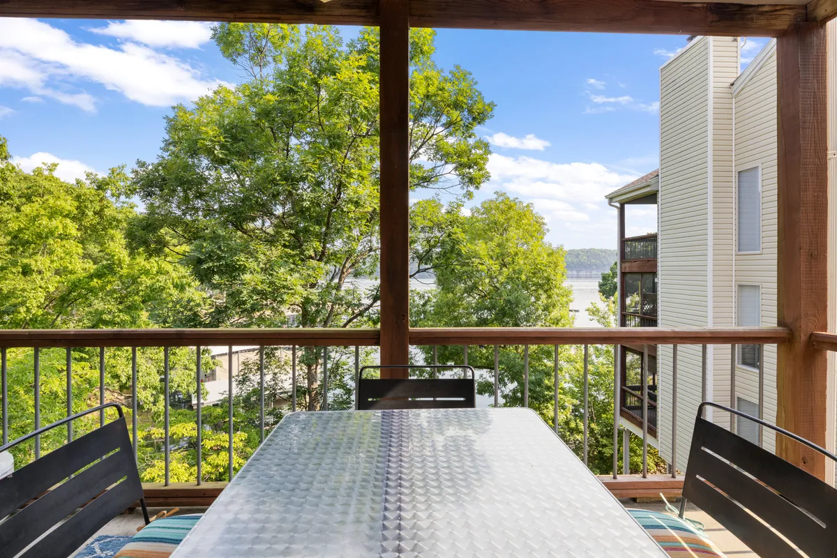 This image captures an outdoor dining table set beneath lush green trees, with a tranquil water view in the background. The setting suggests an inviting space for morning coffee, romantic dinners, or simply taking in the scenery together.