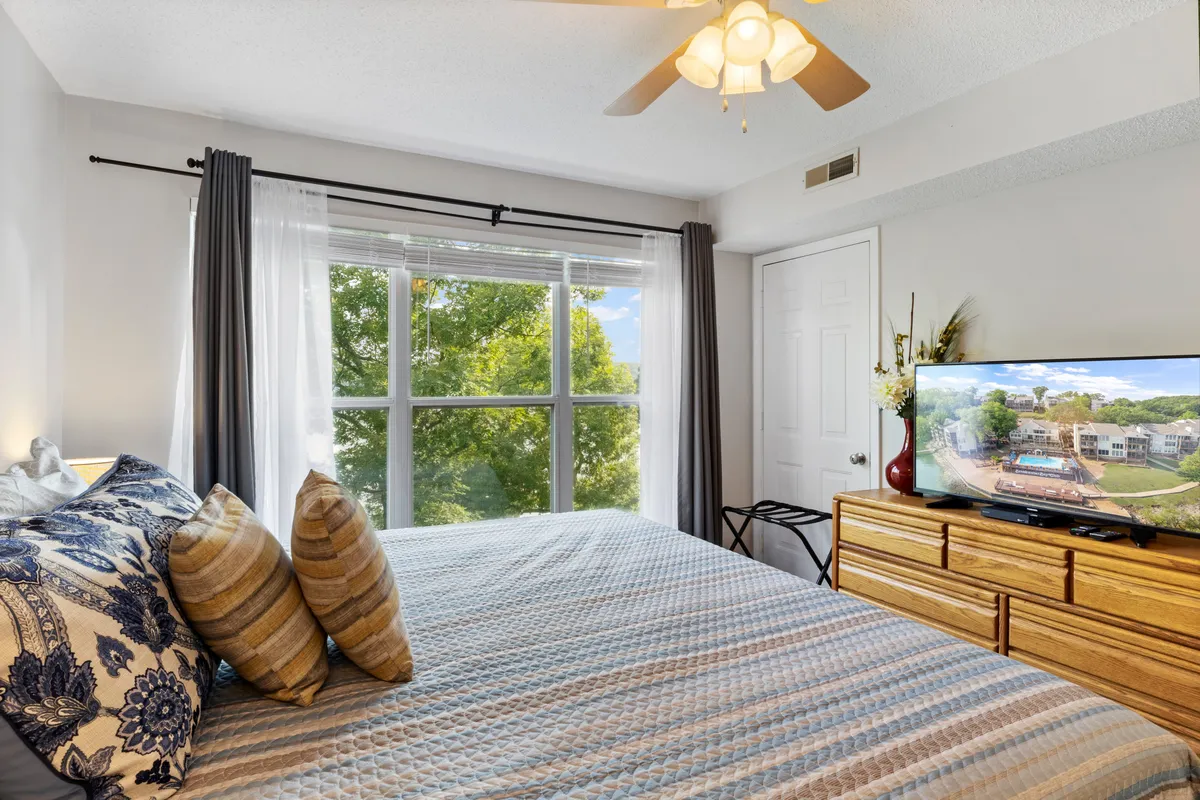 A cozy bedroom featuring a plush bed adorned with decorative pillows, a large window letting in ample daylight, and a television placed on a dresser. The room’s calm color palette and soft furnishings create a soothing atmosphere ideal for unwinding after a busy day.