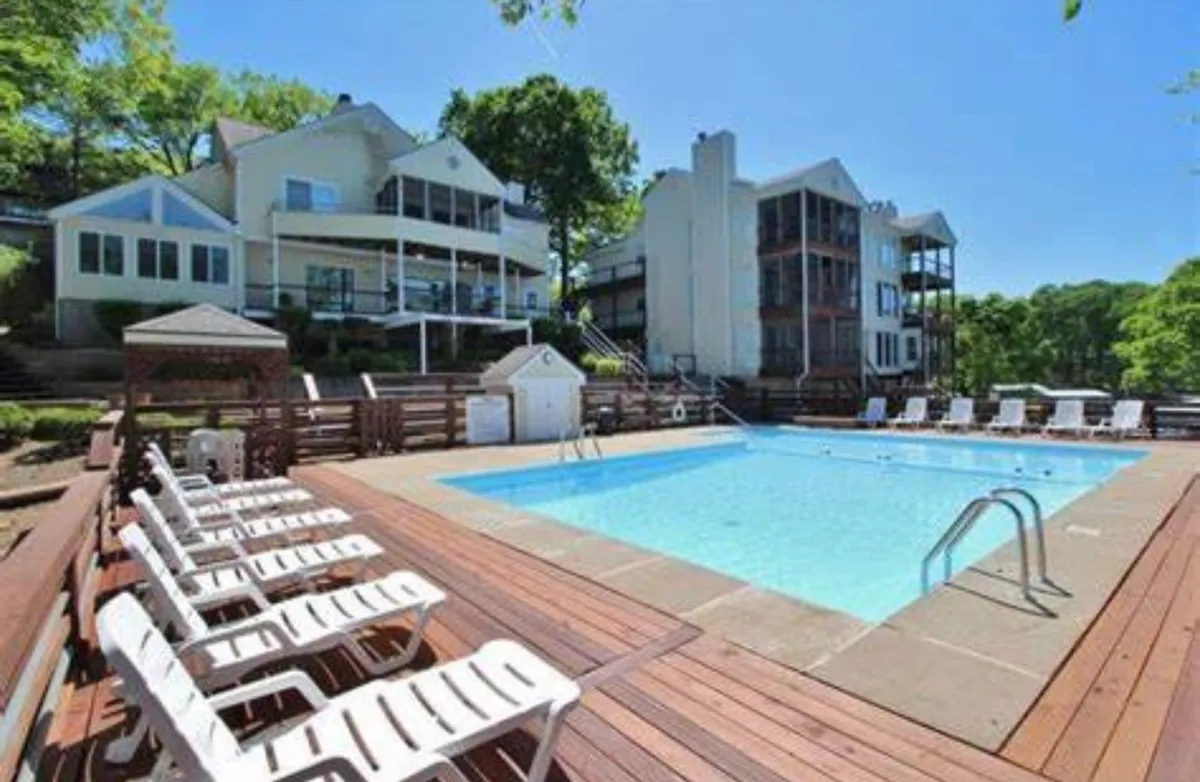 A vibrant exterior view of the community swimming pool surrounded by lounge chairs and shaded by mature trees, with multi-story houses in the background. The setting is bright and lively, ideal for swimming, sunbathing, and social gatherings—especially during the summer season.